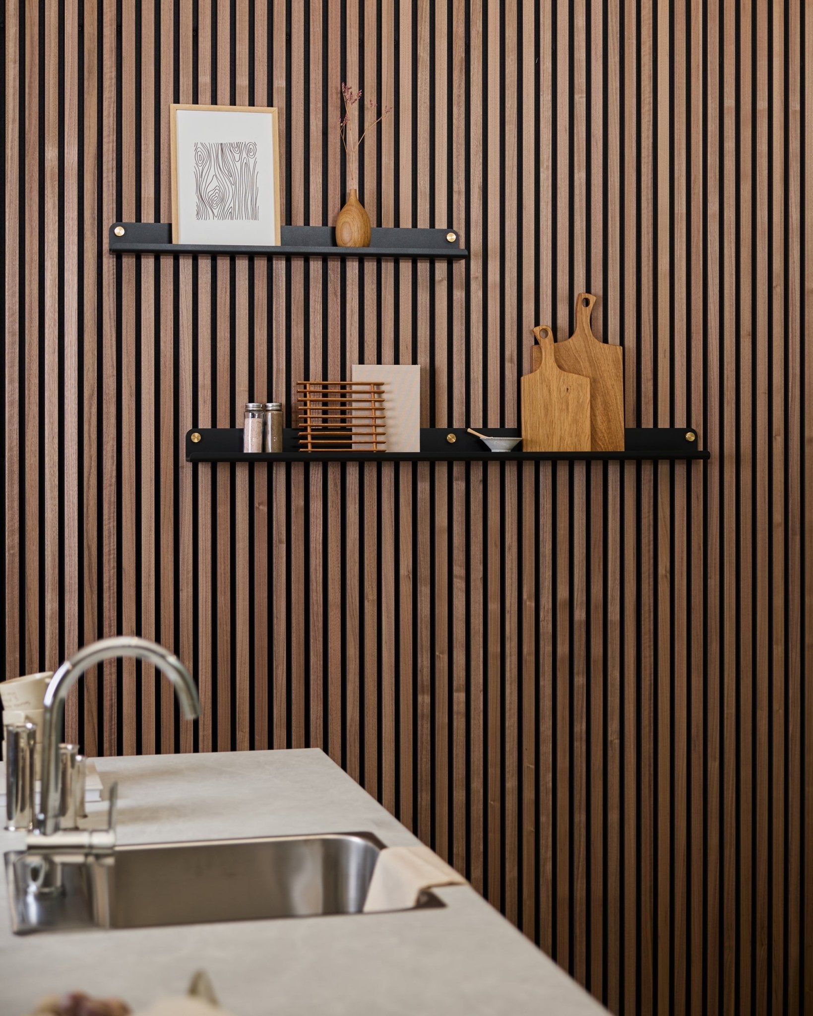 Minimalistische Küche mit einer vertical Akupanel Akustikwand aus Holzlamellen, black Regalböden, Schneidebrettern, small Deko-Objekten und einer modernen Spüle davor.